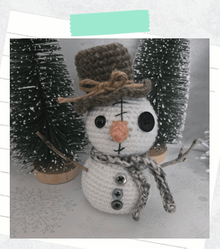 A completed crochet snowman showing all buttons, twig arms, top hat, and carrot nose.